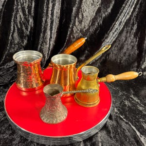 May include: A collection of copper and brass coffee pots with wooden handles, displayed on a red, circular tray. The pots vary in size and design, showcasing a range of metallic finishes and textures. The background is a dark, textured fabric.