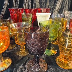 May include: A collection of colorful vintage glassware in shades of red, amber, green, and purple. The glasses vary in shape and design, including goblets and tumblers, with etched and textured patterns. The arrangement suggests a display of antique drinkware.