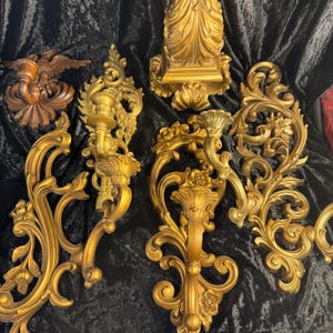 May include: A collection of gold-toned wall sconces and decorative accents featuring intricate scrollwork, floral motifs, and acanthus leaf designs. One dark brown wall sconce depicts an eagle figure. The pieces exhibit ornate, curved, and symmetrical patterns.