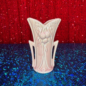 May include: A pale pink ceramic vase with a stylized floral design. The vase features a flared top with a central floral motif and two side handles. The vase is approximately 8 inches tall.