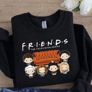 以下が含まれることがあります： 「FRIENDS」の文字とカラフルなドット、そして「THE TELEVISION SERIES」の文字が白でプリントされた黒いスウェットシャツ。その下に、番組のキャラクターとオレンジ色のソファの漫画イラストが描かれています。