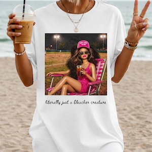 May include: White t-shirt featuring a graphic of a doll in a pink dress and hat, seated in a pink chair, holding a drink. The text "literally just a bleacher creature" is printed below the graphic.