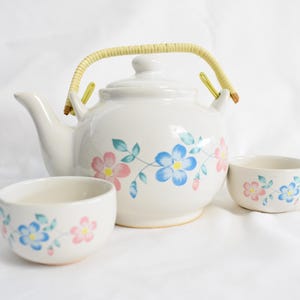 May include: A white ceramic teapot with a floral design in pink, blue, and green. The teapot has a woven handle and comes with two matching teacups.