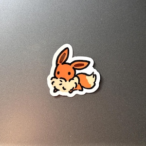 May include: A sticker depicting a cartoon fox-like character with orange and white fur, brown ears, and a fluffy neck. The sticker has a white border against a grey background.