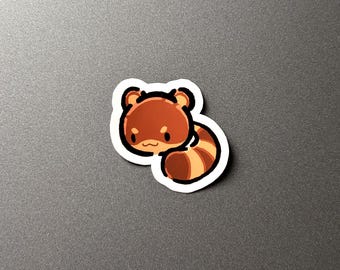 May include: A sticker featuring a cartoon red panda with a white border. The red panda has brown fur with lighter brown and tan stripes on its tail. The sticker is on a gray surface.