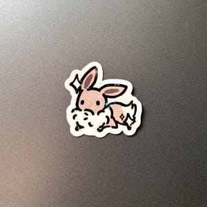 May include: A sticker of a cartoon bunny with brown ears and body, and a white fluffy belly. The sticker has a white border and a glittery, holographic effect. The bunny is depicted in a running pose.