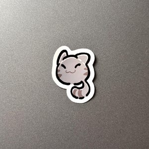 以下が含まれることがあります： 笑顔の漫画の猫のステッカー。猫は灰色の縞模様と黒いアウトラインです。ステッカーは灰色の表面にあります。