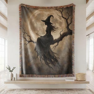 May include: A woven throw blanket featuring a dark silhouette of a witch in a pointed hat sitting on a gnarled tree branch against a large, glowing full moon. The blanket has a fringed edge with multi-colored threads and a sepia-toned color palette.