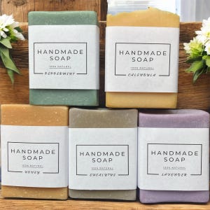 May include: A selection of handmade soaps in shades of green, yellow, and purple. Each bar is wrapped with a white label that reads "HANDMADE SOAP" and lists the scent: Peppermint, Calendula, Honey, Eucalyptus, and Lavender. The soaps are labeled as 100% natural.