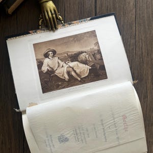 Puede incluir: Un libro abierto con una impresi&oacute;n en tonos sepia de un hombre con sombrero, con el texto "GOETHE IN ITALIEN". Un marcap&aacute;ginas dorado en forma de mano con un anillo sujeta las p&aacute;ginas abiertas. El libro est&aacute; sobre una superficie de madera.