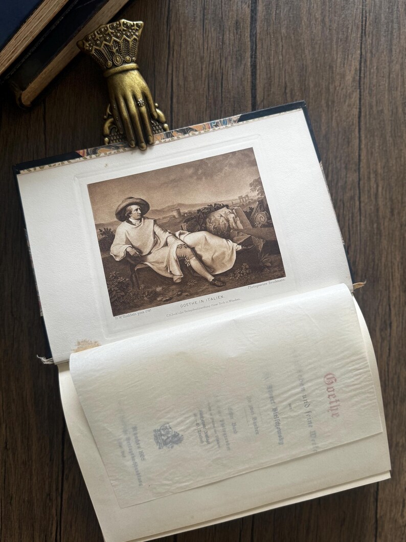 Puede incluir: Un libro abierto con una impresi&oacute;n en tonos sepia de un hombre con sombrero, con el texto "GOETHE IN ITALIEN". Un marcap&aacute;ginas dorado en forma de mano con un anillo sujeta las p&aacute;ginas abiertas. El libro est&aacute; sobre una superficie de madera.