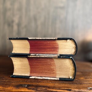Puede incluir: Dos libros antiguos apilados. Tienen cubiertas azul oscuro con letras doradas y p&aacute;ginas envejecidas de color marr&oacute;n rojizo. Los bordes de las p&aacute;ginas son de color amarillo dorado. Los libros descansan sobre una superficie de madera.