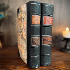 Puede incluir: Dos libros antiguos con cubiertas verde oscuro y etiquetas decorativas. Las etiquetas del lomo dicen "Goethe" y tienen los n&uacute;meros "1" y "2". El libro de la izquierda tiene un dise&ntilde;o marmolado colorido en su cubierta. Los libros se exhiben sobre una superficie de madera.