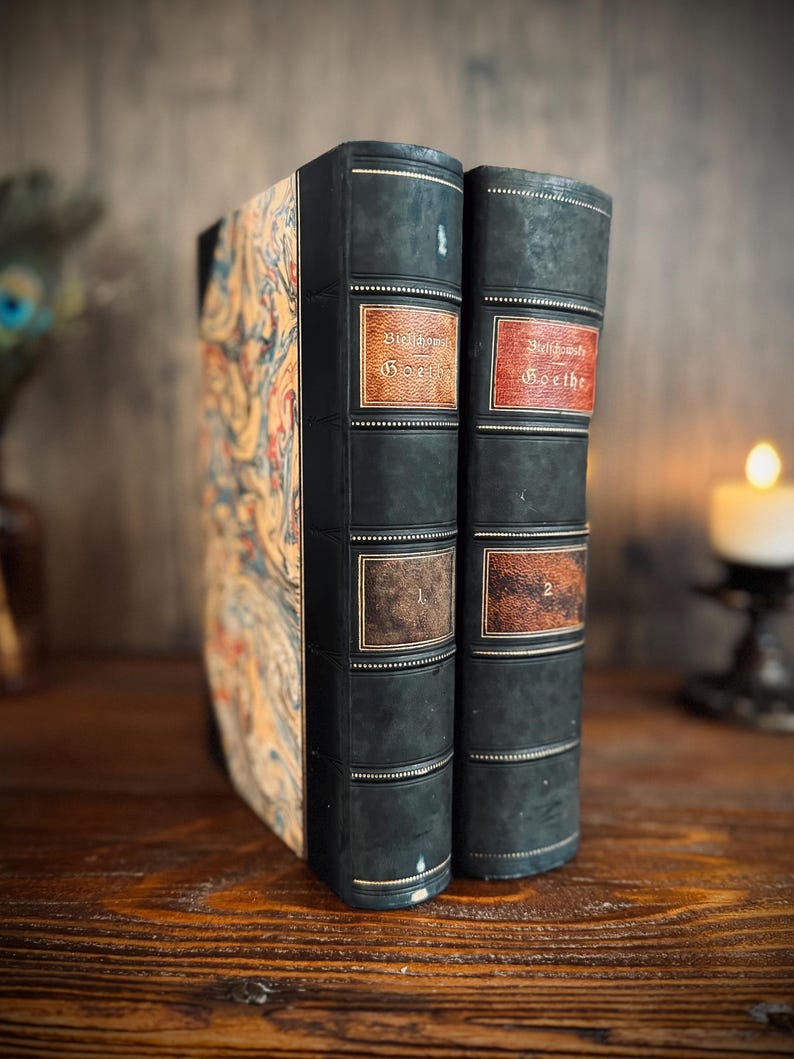 Puede incluir: Dos libros antiguos con cubiertas verde oscuro y etiquetas decorativas. Las etiquetas del lomo dicen "Goethe" y tienen los n&uacute;meros "1" y "2". El libro de la izquierda tiene un dise&ntilde;o marmolado colorido en su cubierta. Los libros se exhiben sobre una superficie de madera.