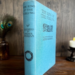 May include: A light blue hardcover book titled The Ring of the Niblung, illustrated by Arthur Rackham. The cover features black text and a dragon illustration. The spine displays the title, illustrator name, a circular serpent graphic, and Heinemann.
