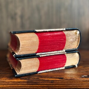 Puede incluir: Dos libros antiguos apilados sobre una superficie de madera. Los libros tienen lomos rojos, p&aacute;ginas doradas y cubiertas negras. Las p&aacute;ginas envejecidas muestran signos de desgaste, lo que sugiere que son vintage o muy le&iacute;dos.