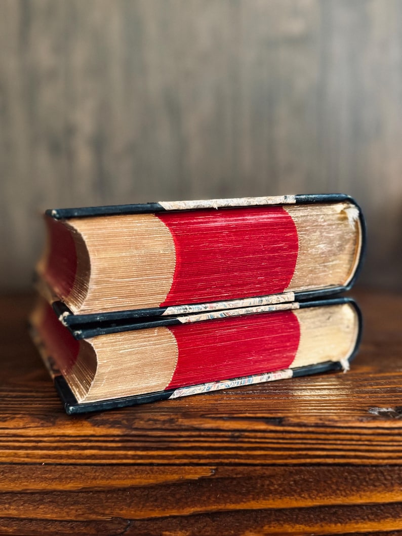 Puede incluir: Dos libros antiguos apilados sobre una superficie de madera. Los libros tienen lomos rojos, p&aacute;ginas doradas y cubiertas negras. Las p&aacute;ginas envejecidas muestran signos de desgaste, lo que sugiere que son vintage o muy le&iacute;dos.
