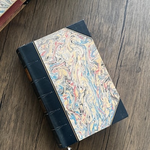 Puede incluir: Un libro antiguo con una cubierta veteada en tonos azules, rojos y amarillos. El lomo es azul oscuro y el libro descansa sobre una superficie de madera. Los bordes del libro son visibles, lo que sugiere una colecci&oacute;n de literatura antigua.
