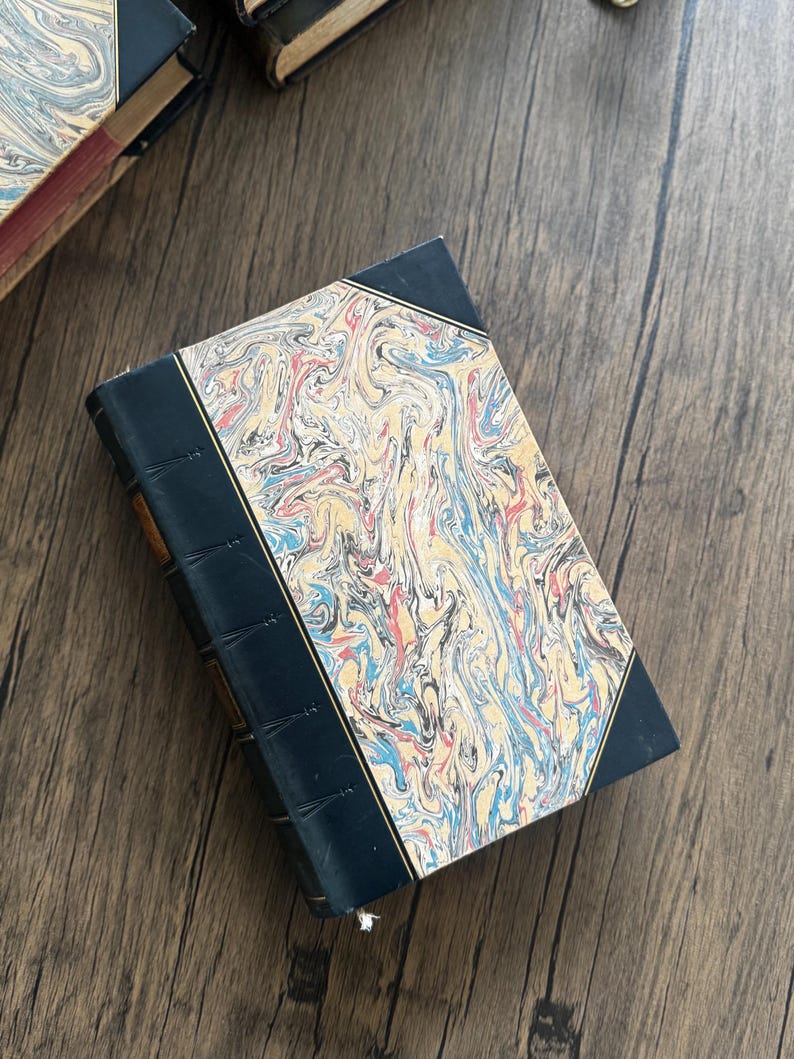 Puede incluir: Un libro antiguo con una cubierta veteada en tonos azules, rojos y amarillos. El lomo es azul oscuro y el libro descansa sobre una superficie de madera. Los bordes del libro son visibles, lo que sugiere una colecci&oacute;n de literatura antigua.