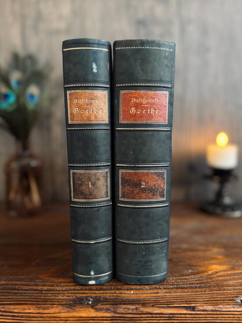 Puede incluir: Dos libros antiguos con cubiertas verde oscuro y letras doradas. Las lomos presentan etiquetas que dicen "Bielschowsky Goethe" y los n&uacute;meros "1" y "2". Los libros se exhiben sobre una superficie de madera, con un fondo borroso.