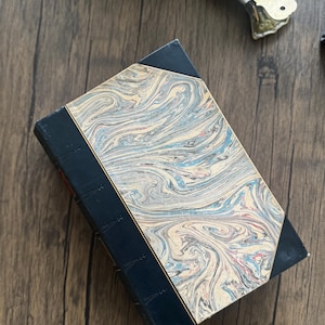 Puede incluir: Libro antiguo con una cubierta veteada en tonos azules, crema y rojo. El lomo es azul oscuro y los bordes de la cubierta est&aacute;n angulados. El libro est&aacute; sobre una superficie de madera.