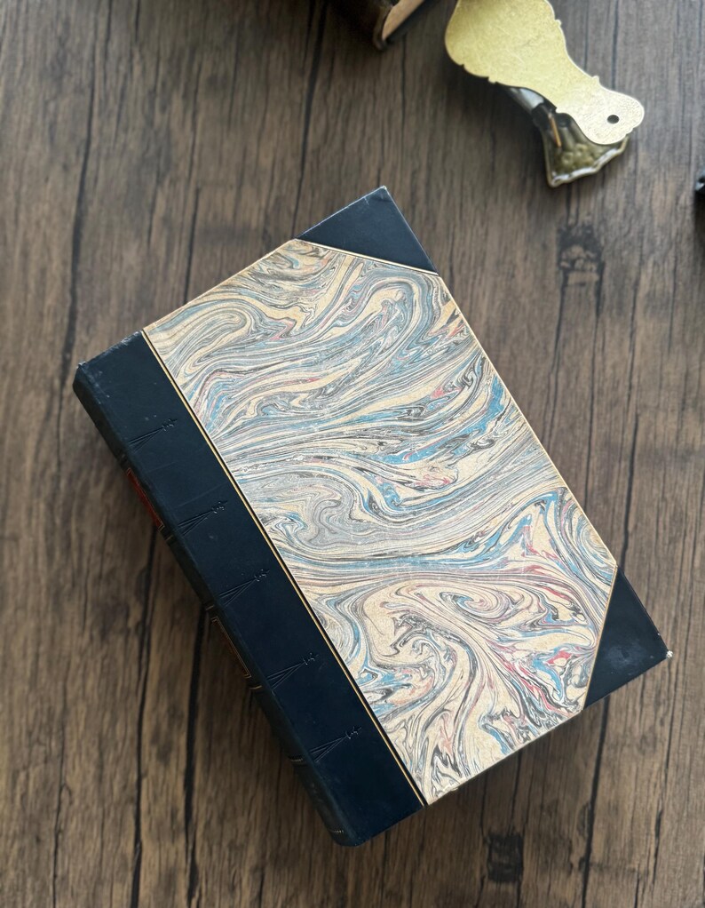 Puede incluir: Libro antiguo con una cubierta veteada en tonos azules, crema y rojo. El lomo es azul oscuro y los bordes de la cubierta est&aacute;n angulados. El libro est&aacute; sobre una superficie de madera.