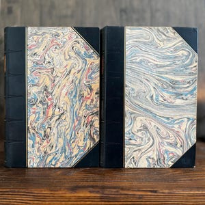 Puede incluir: Dos libros antiguos con cubiertas de papel veteado. Las cubiertas presentan un patr&oacute;n arremolinado de azul, rojo, amarillo y blanco. Las lomos son negros con detalles dorados. Los libros est&aacute;n sobre una superficie de madera.