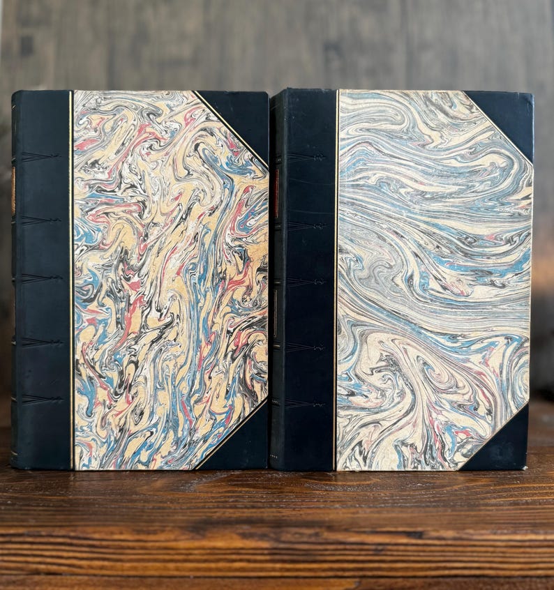 Puede incluir: Dos libros antiguos con cubiertas de papel veteado. Las cubiertas presentan un patr&oacute;n arremolinado de azul, rojo, amarillo y blanco. Las lomos son negros con detalles dorados. Los libros est&aacute;n sobre una superficie de madera.