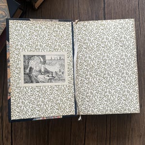 Puede incluir: Un libro abierto con una ilustraci&oacute;n y texto detallados. Las p&aacute;ginas interiores presentan un patr&oacute;n de hojas verdes repetido sobre un fondo crema. La ilustraci&oacute;n es un dibujo en blanco y negro de una persona en una mesa.