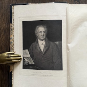Puede incluir: Un libro abierto con un retrato en blanco y negro de un hombre sosteniendo un papel. El retrato est&aacute; enmarcado dentro de las p&aacute;ginas del libro. La columna del libro tiene un dise&ntilde;o.