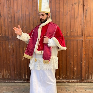 May include: A religious costume featuring a white mitre with gold trim, a red and burgundy cape with gold accents, and a long white robe. The costume includes a red sash and red boots. The outfit is designed for a religious figure.
