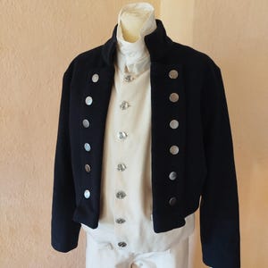 Puede incluir: Una chaqueta azul marino oscuro sobre un chaleco y pantalones de color crema. La chaqueta presenta dos filas verticales de botones plateados. El chaleco crema también tiene una fila vertical de botones plateados más pequeños y detallados.