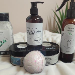 May include: A collection of personal care products including a square bottle labeled Variant Eucalyptus Mint, a pump bottle labeled Head to Toe Hair/Body Wash, a spray bottle labeled Simply Bamboo, two black jars labeled Body Scrub, and a round bath bomb.