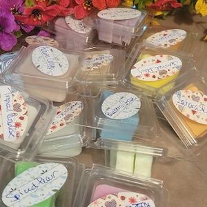 May include: An assortment of scented wax melts in clear plastic containers. The melts are various colors, including pink, green, yellow, and blue. Each container has a label with a handwritten scent name, such as "Rose Bouquet" and "Spiced Pear". Colorful flowers are in the background.