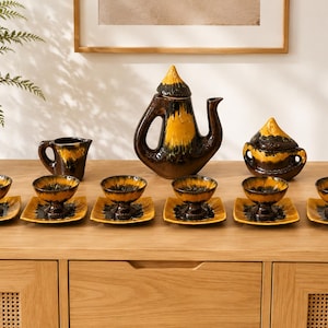 May include: A ceramic tea set featuring a tall teapot with a lid, a creamer, a lidded sugar bowl, and six footed cups on square saucers. The pieces have a glossy finish with a dark brown base and a vibrant yellow glaze dripping from the top.