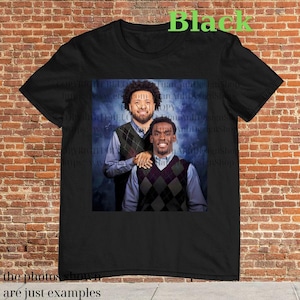 Könnte beinhalten: Schwarzes T-Shirt mit einem Foto von zwei Männern, die Pullover tragen. Die Männer lächeln und stehen nebeneinander. Der Hintergrund ist ein blaues und weißes abstraktes Design.