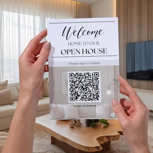 May include: A sign that reads "Welcome Home to Our Open House" is held up in front of a living room. The sign has a QR code and the words "Scan to Sign In" below it. The sign is white with black text.