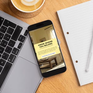 May include: A smartphone displays the text "GROW WHERE YOU BELONG" on a yellow background. A laptop, a cup of coffee, a pen, and a notepad are on a wooden desk. The laptop keyboard is visible.