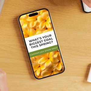 May include: A smartphone displays a floral background with the text "WHAT'S YOUR BIGGEST GOAL THIS SPRING?" and "Comment your answer below." The phone is on a wooden surface, with a notebook, eraser, and other items nearby.