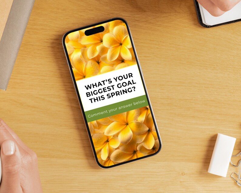 May include: A smartphone displays a floral background with the text "WHAT'S YOUR BIGGEST GOAL THIS SPRING?" and "Comment your answer below." The phone is on a wooden surface, with a notebook, eraser, and other items nearby.