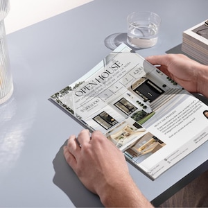 May include: A person holding an open house flyer with the text "OPEN HOUSE" and property details. The flyer features images of a modern home, with details including price, number of bedrooms, bathrooms, and square footage. A glass of water and a decorative jar are also visible.