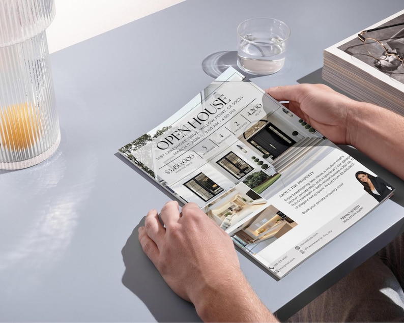 May include: A person holding an open house flyer with the text "OPEN HOUSE" and property details. The flyer features images of a modern home, with details including price, number of bedrooms, bathrooms, and square footage. A glass of water and a decorative jar are also visible.