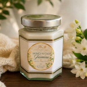 Pode incluir: Uma vela em pote de vidro hexagonal com tampa prateada. O rótulo diz "JK Enchanted Flames Honeysuckle Jasmine" com detalhes florais. A vela é cercada por flores brancas e vegetação.
