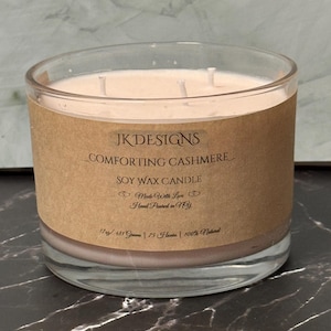 Puede incluir: Una vela de vidrio transparente en un frasco con una etiqueta marrón que dice "JK DESIGNS _COMFORTING CASHMERE_ SOY WAX CANDLE Made With Love Hand Poured in NY 17 oz / 481 Grams | 75 Hours | 100% Natural"