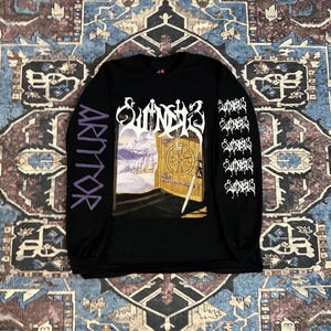 Może przedstawiać: Czarna koszulka z długim rękawem z graficznym wzorem. Wzór przedstawia drzwi, miecz i słowo "Sunder" w kolorze białym. Lewy rękaw ma słowo "Arttor" w kolorze fioletowym, a prawy rękaw ma słowo "Sunder" powtórzone w kolorze białym.