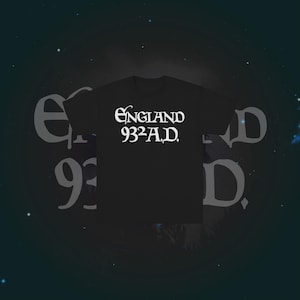 Pode incluir: Uma t-shirt preta de manga curta com texto branco impresso no peito. O texto diz "ENGLAND 932 A.D." numa fonte decorativa estilizada. A peça tem gola redonda e um corte padrão, apresentada contra um fundo escuro.
