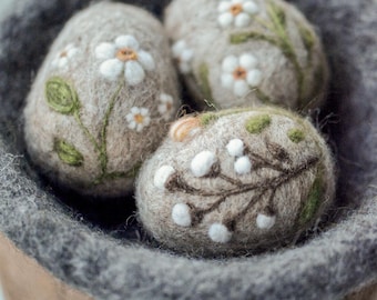 May include: Three handcrafted, felted eggs with floral embroidery, nestled in a gray felt basket. The eggs are beige with white daisy and green leaf designs. The basket has a brown base and gray rim.