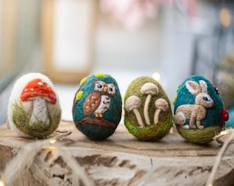 May include: Four decorative Easter eggs with felted designs. One egg features a red and white mushroom, another an owl, a third with mushrooms, and the last with a rabbit. The eggs are displayed on a wooden surface.