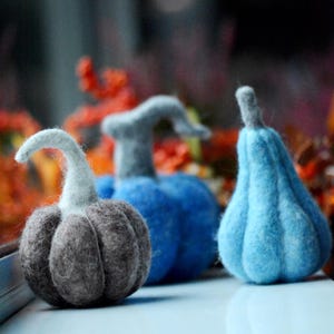 May include: Three felt pumpkins in shades of gray and blue. The pumpkins have textured surfaces and curved stems. The pumpkins are arranged on a white surface, with a blurred background of fall foliage.