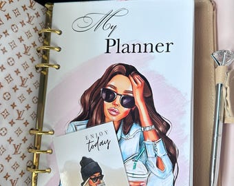 May include: A gold and black planner with a pink background and a woman with sunglasses and a blue shirt. The planner has the text "My Planner" on the cover and a sticker that says "Enjoy today".